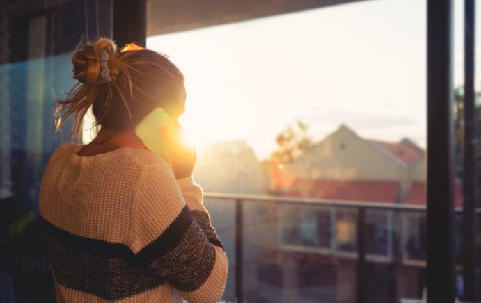 Woman looking through the window at sunset, talking on her mobile phone